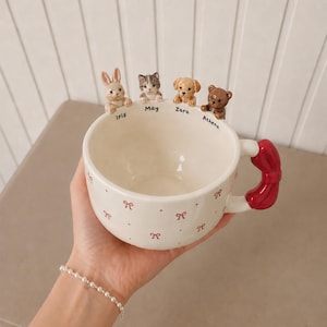 Op de afbeelding: Een crèmekleurige keramische mok met een rood strikhandvat en een patroon van kleine rode strikjes en stippen. De rand is versierd met vier kleine dierenfiguurtjes: een konijn genaamd Iris, een kat May, een hond Zara en een beer genaamd Athena.