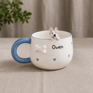Puede incluir: Una taza de cerámica blanca con asa y borde azules, con una figura de perro husky en 3D. La taza tiene un adorno en forma de hueso y detalles de corazones azules. El nombre "Owen" está impreso en la taza.