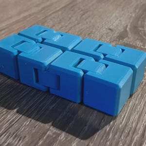 May include: A blue fidget toy, composed of connected rectangular blocks. The toy is designed to bend and flex, offering a tactile experience. The blocks are a solid, light blue color, and the toy is resting on a wooden surface.