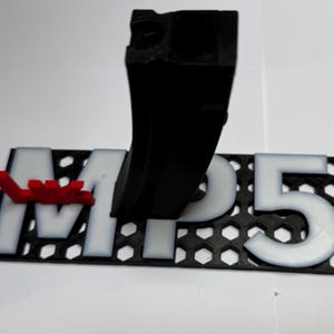 May include: A black magazine stand featuring a white MP5 label on a black hexagonal grid base with a small red accent piece. The stand is designed to hold a black firearm magazine in an upright position, displaying the model name clearly.