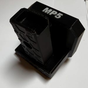 May include: A black 3D-printed mount featuring a rectangular base and a vertical, hollow, hexagonal-patterned support column. The top surface of the mount displays the white text MP5, indicating its specific compatibility or model designation.
