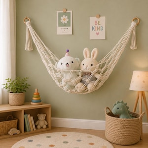 May include: A cream-colored macrame toy hammock suspended by two wooden rings, holding a white plush bear wearing a party hat and a beige plush rabbit. The hammock features a knotted net design with decorative tassels hanging from each side.