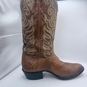 Könnte beinhalten: Ein brauner Cowboystiefel aus Leder mit einem strukturierten Unterteil in Schlangenlederoptik und einem hohen Schaft, der mit aufwendigen, hellen Ziernähten versehen ist. Der Stiefel verfügt über einen Blockabsatz und eine spitz zulaufende Form.
