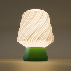 May include: A modern table lamp with a white, swirling shade and a bright green base. The shade has a textured, layered design, and the lamp emits a soft, warm glow. The lamp is a decorative lighting fixture.