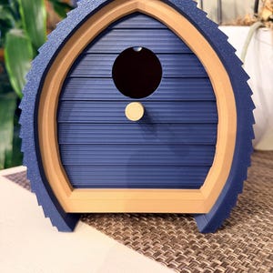 May include: A blue and tan birdhouse with a round entry hole and a small wooden perch. The birdhouse has a textured roof and a layered design, with a light brown frame and a dark blue body. The birdhouse is sitting on a brown surface.