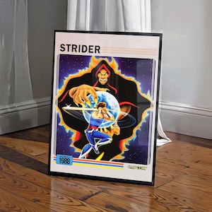 May include: A framed poster featuring the Strider video game artwork. The graphic displays a central character wielding a sword against a large, dark, hooded figure with yellow claws. The text reads STRIDER, NOVEMBER 1988, and CAPCOM.