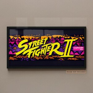 May include: A rectangular wall art print featuring the yellow Street Fighter II logo centered over a dark background with orange and purple abstract patterns. A small pink Capcom USA logo is on the right. Text below reads: FRAME NOT INCLUDED.