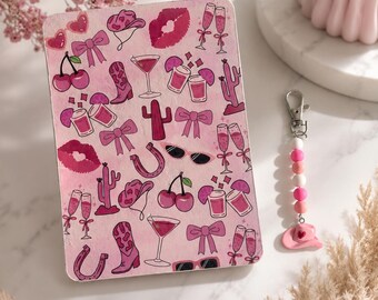 Könnte beinhalten: Ein rechteckiges Notizbuch mit rosa Hintergrund und einem wiederkehrenden Muster aus Cowboyhüten, Stiefeln, Schleifen, Kirschen, Kakteen, Hufeisen, Lippen, Sonnenbrillen und Cocktailgläsern, dazu ein Perlen-Schlüsselanhänger mit einem rosa Cowboyhut.