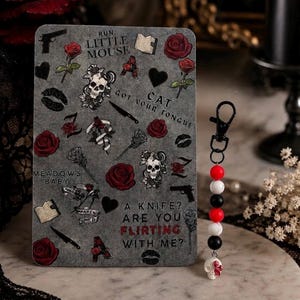 Op de afbeelding: Een rechthoekige grijze kaart met illustraties van rode rozen, schedels, messen, boeken en lippen, met de tekst RUN LITTLE MOUSE, CAT GOT YOUR TONGUE, MEADOWS BABY en A KNIFE? ARE YOU FLIRTING WITH ME? Daarnaast ligt een sleutelhanger met kralen.