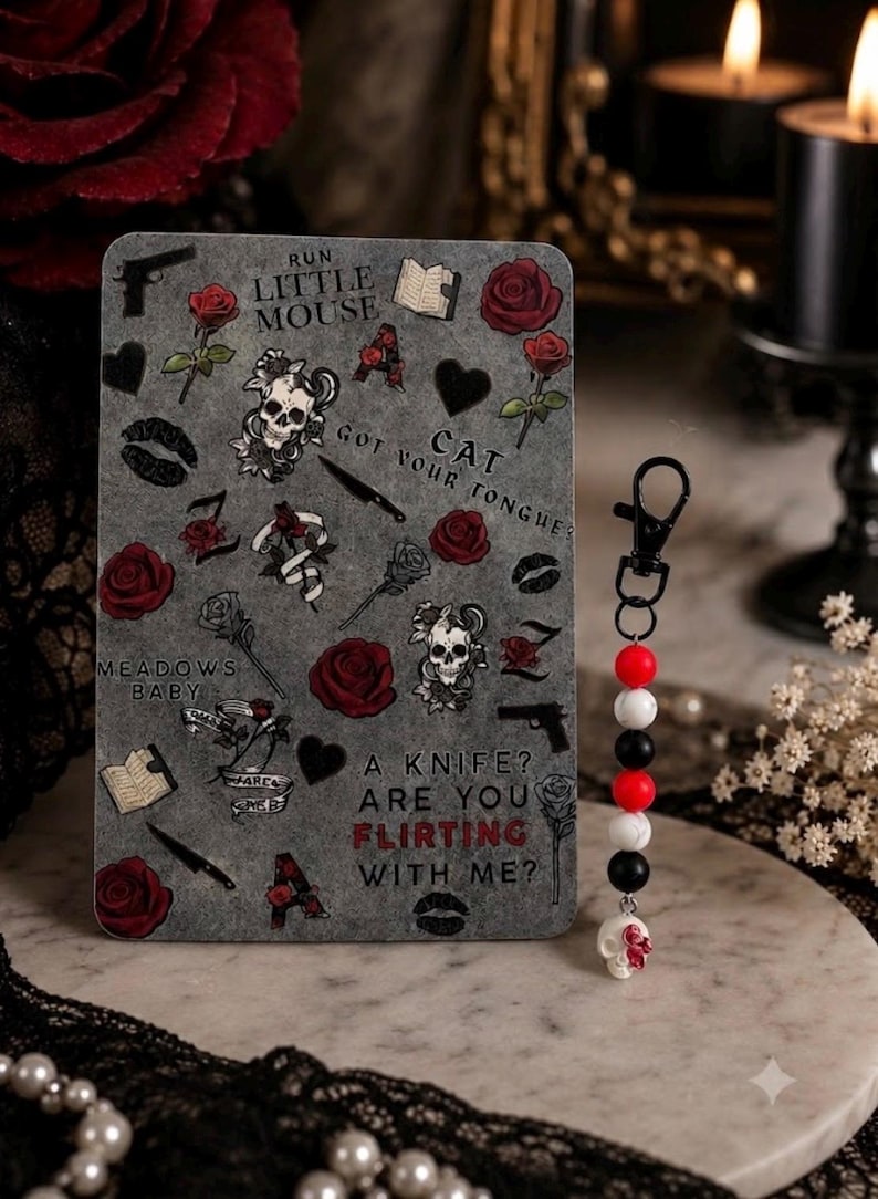 Op de afbeelding: Een rechthoekige grijze kaart met illustraties van rode rozen, schedels, messen, boeken en lippen, met de tekst RUN LITTLE MOUSE, CAT GOT YOUR TONGUE, MEADOWS BABY en A KNIFE? ARE YOU FLIRTING WITH ME? Daarnaast ligt een sleutelhanger met kralen.