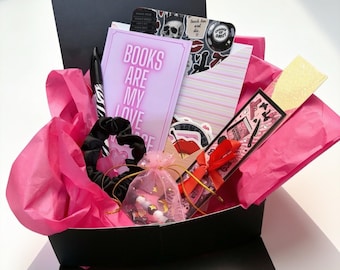 Könnte beinhalten: Eine schwarze Geschenkbox gefüllt mit leuchtend rosa Seidenpapier, die eine Karte mit dem Text BOOKS ARE MY LOVE LANGUAGE, ein schwarzes Haargummi, einen kleinen rosa Organzabeutel mit Perlen, ein Lesezeichen mit rotem Band und Aufkleber enthält.