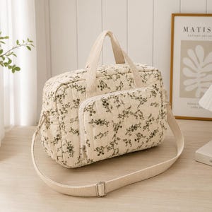 Peut inclure: Un sac rectangulaire matelassé de couleur crème orné d'un petit motif floral vert foncé. Le sac comprend deux poignées supérieures, une poche frontale zippée et une bandoulière beige réglable avec des boucles métalliques.