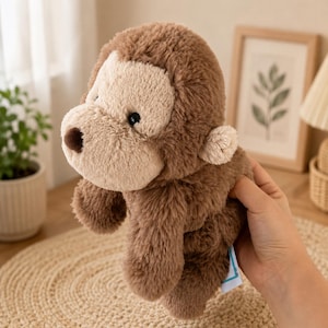 May include: A plush monkey toy featuring soft, shaggy brown fur on the body and head, with a lighter beige face, small rounded ears, and a dark brown nose. The stuffed animal has black bead eyes and a small white fabric tag attached to the side.