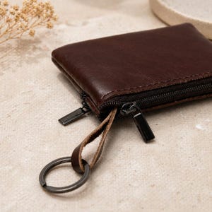 May include: A dark brown leather coin purse with a black zipper and a key ring. The purse is rectangular and has a smooth texture. The zipper pulls are rectangular and black.