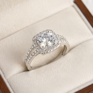 Peut inclure: Une bague argentée dotée d'une grande pierre centrale ronde et transparente, entourée d'un halo carré de petites pierres transparentes. L'anneau est divisé en deux rangées, chacune incrustée de petites pierres transparentes scintillantes.