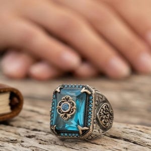Peut inclure: Bague en argent ornée d'une grande pierre précieuse rectangulaire bleu clair. La bague présente des détails complexes autour de la pierre et sur le bandeau, avec de petites pierres bleues sur les bords. Posée sur une surface en bois.