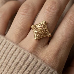 Peut inclure: Une bague dorée avec un design ajouré orné. La bague présente une pièce centrale de forme carrée avec des motifs de filigrane complexes. La bague est portée sur un doigt, sur un fond beige neutre.