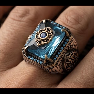 Peut inclure: Bague de style antique avec une grande pierre précieuse rectangulaire bleu clair. La monture dorée est ornée de motifs complexes et de petites pierres turquoise. Une petite pierre bleue est sertie au centre.