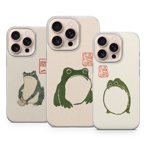 May include: Three phone cases with a beige background and green frog illustrations. The cases feature different frog designs, including one with a bowl and text, and another with a red stamp. The cases are designed to fit a smartphone.