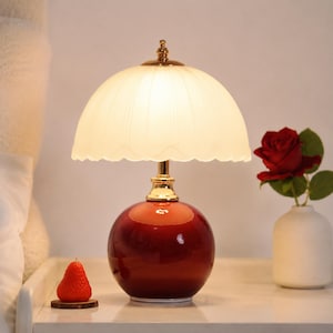 Puede incluir: Lámpara de mesa de cerámica roja con base redonda y pantalla festoneada de color crema. La lámpara tiene detalles dorados. Una rosa roja en un jarrón blanco está junto a la lámpara, sobre una superficie blanca.