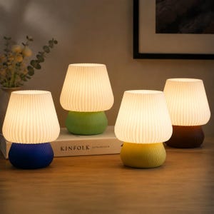 May include: Four mushroom-shaped table lamps with pleated white shades and colorful bases. The bases are blue, green, yellow, and brown. The lamps are arranged on a wooden surface, with a book and a framed picture in the background.
