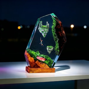 May include: A unique resin sculpture featuring a miniature fantasy landscape with floating islands, a castle, and tiny figures. The sculpture is encased in a clear, faceted resin block, mounted on a wooden base. The landscape includes green moss and a red and brown rock formation.