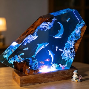 May include: A decorative resin lamp depicting an underwater scene with dolphins, jellyfish, and coral. The lamp has a wooden base and a clear resin body with embedded sea life, illuminated by blue LED lights. The lamp's dimensions are approximately 20 cm tall and 15 cm wide.