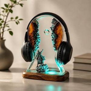 Op de afbeelding: Een decoratieve hoofdtelefoonstandaard met een houten basis, een helder acryl frame en een gedetailleerde fantasiefiguur. De standaard heeft een blauwe gloed en houdt een zwarte hoofdtelefoon vast. Het ontwerp bevat oranje en teal accenten.