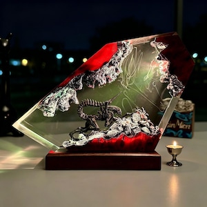 May include: A clear resin sculpture featuring a detailed Alien creature design with a red and white background. The sculpture is mounted on a dark wooden base. A small candle in a silver holder sits nearby.
