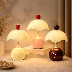 May include: Three decorative table lamps with a retro design. Each lamp features a round base in yellow, pink, and burgundy, topped with a cream-colored, scalloped shade. A red or dark brown sphere sits atop each shade. The lamps are approximately 12 inches tall.