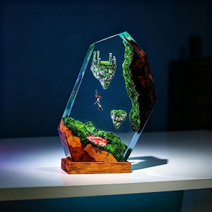 May include: A unique resin art piece featuring a miniature landscape with floating islands, lush green moss, and a wooden base. The clear resin encases the scene, creating a three-dimensional effect. The artwork includes tiny figures, adding a sense of scale and wonder.