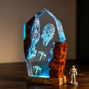 May include: A Star Wars-themed lamp featuring the Millennium Falcon and X-wing fighters encased in a clear resin block. The lamp has a wooden base and a blue light. A Stormtrooper figurine stands nearby.