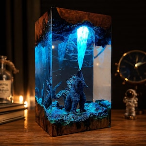 May include: A rectangular resin art piece featuring a blue Godzilla figure emitting a bright blue beam. The sculpture is encased in clear resin with a wooden base and top. The piece is illuminated with blue light, creating a glowing effect.