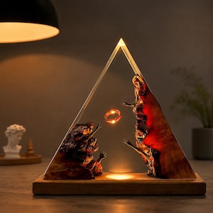 May include: A decorative lamp featuring a clear, triangular resin structure with a wooden base. Inside, figures reach for a glowing ring. The resin is filled with textured, reddish-brown elements, and the lamp emits a warm light.