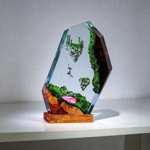 May include: A clear resin sculpture featuring a miniature fantasy landscape. The artwork includes floating islands with miniature castles, lush green foliage, and a small figure. The base is made of wood, and the sculpture is set against a light background.