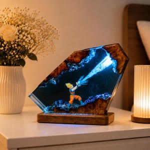 Op de afbeelding: Een decoratieve harslamp met een personage in een oranje en blauwe outfit, die een blauwe energie-explosie uitzendt. De lamp heeft een houten voet en een uniek, schuin ontwerp met bruine en blauwe accenten.