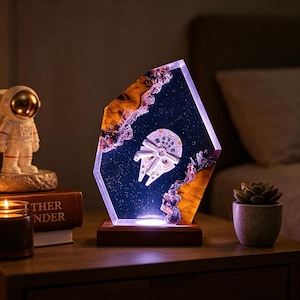 May include: A decorative lamp featuring a spaceship design encased in a clear, geometric resin. The lamp has a wooden base and is illuminated with a soft, warm light. The spaceship is white and gray, set against a dark blue background with a starry effect.