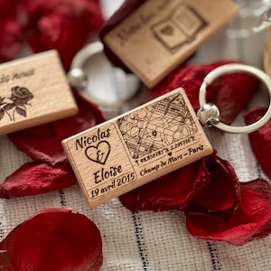 Op de afbeelding: Houten sleutelhangers met gegraveerde ontwerpen. Eén sleutelhanger toont de namen "Nicolas & Eloise", een hartsymbool, de datum "19 april 2015" en coördinaten voor Champ de Mars-Paris. Andere sleutelhangers hebben kaart- en bloemmotieven.