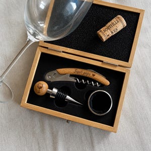 Op de afbeelding: Een houten wijngeschenkset met een kurkentrekker, flesstopper en een wijnflesring. De kurkentrekkers handvat is gegraveerd met "Super papa". De set bevat een kurk met de tekst "BOUTEILLE A LA".