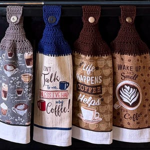 May include: Four hanging kitchen towels with crocheted tops and buttons. The towels feature coffee-themed patterns and text including "Don't talk to me unless I've had my coffee!", "Life happens coffee helps", and "Wake up and smell the coffee".