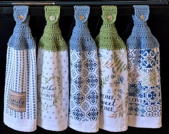 May include: Five hanging kitchen towels with crocheted tops in blue and green, each secured with a button. The towels feature various prints including vertical stripes, floral wreaths with text, geometric tile patterns, and botanical motifs.