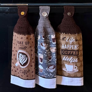 May include: Three hanging kitchen towels with crocheted tops and buttons. The left towel is brown with coffee beans and text reading WAKE UP AND SMELL THE COFFEE. The center is gray with coffee cup graphics. The right is brown with LIFE HAPPENS COFFEE HELPS.