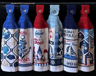 May include: Six hanging kitchen towels with nautical themes. Each towel has a crocheted top in blue or red, with a button closure. The towels feature images of seashells, anchors, gnomes, sailboats, and text like "Home where you drop your anchor" and "Sailboat Rentals".