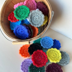 May include: A collection of round, crocheted scrubbers in a variety of vibrant colors including red, blue, green, yellow, orange, purple, black, and gray. Some scrubbers are contained within a round wooden basket, while others are scattered below.
