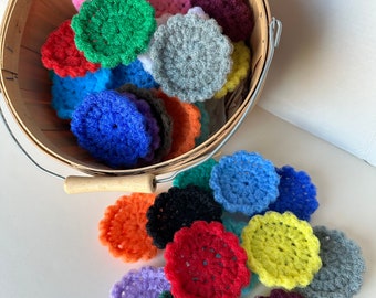 May include: A collection of round, crocheted scrubbers in a variety of vibrant colors including red, blue, green, yellow, orange, purple, black, and gray. Some scrubbers are contained within a round wooden basket, while others are scattered below.