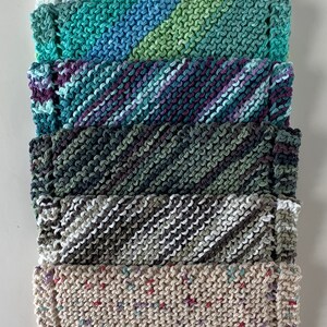 Puede incluir: Una pila de seis pa&ntilde;os de cocina de punto en varios colores y patrones, incluyendo blanco con motas amarillas, rayas verdes y azules, rayas moradas y azules, verde oscuro, rayas grises y blancas, y beige con motas multicolores.