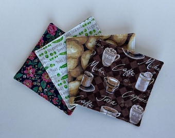 May include: Four fabric coasters with different designs. One coaster features a floral pattern in pink, green, and black. Another coaster has a pattern of green peas in pods on a white background. The third coaster has a pattern of brown potatoes on a white background. The fourth coaster has a brown background with a checkered pattern and a coffee theme with text that reads "Mocha", "Java", "Caffe", and "Cappuccino".