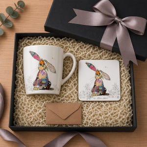 May include: A gift set comprising a white ceramic mug and coaster, each decorated with a colourful patchwork bunny illustration. The items are presented in a black gift box with a grey ribbon, and a small brown envelope.