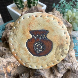 May include: A round, beige ceramic coaster with a brown pot design and a spiral center. The coaster has a series of small holes around the edge. The coaster is displayed on a piece of driftwood.