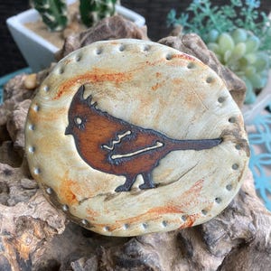 May include: A round, ceramic decorative piece with a bird design. The bird is brown with a dark outline, set against a beige background with orange streaks. The edge has small, evenly spaced holes. The piece is displayed on a piece of driftwood.